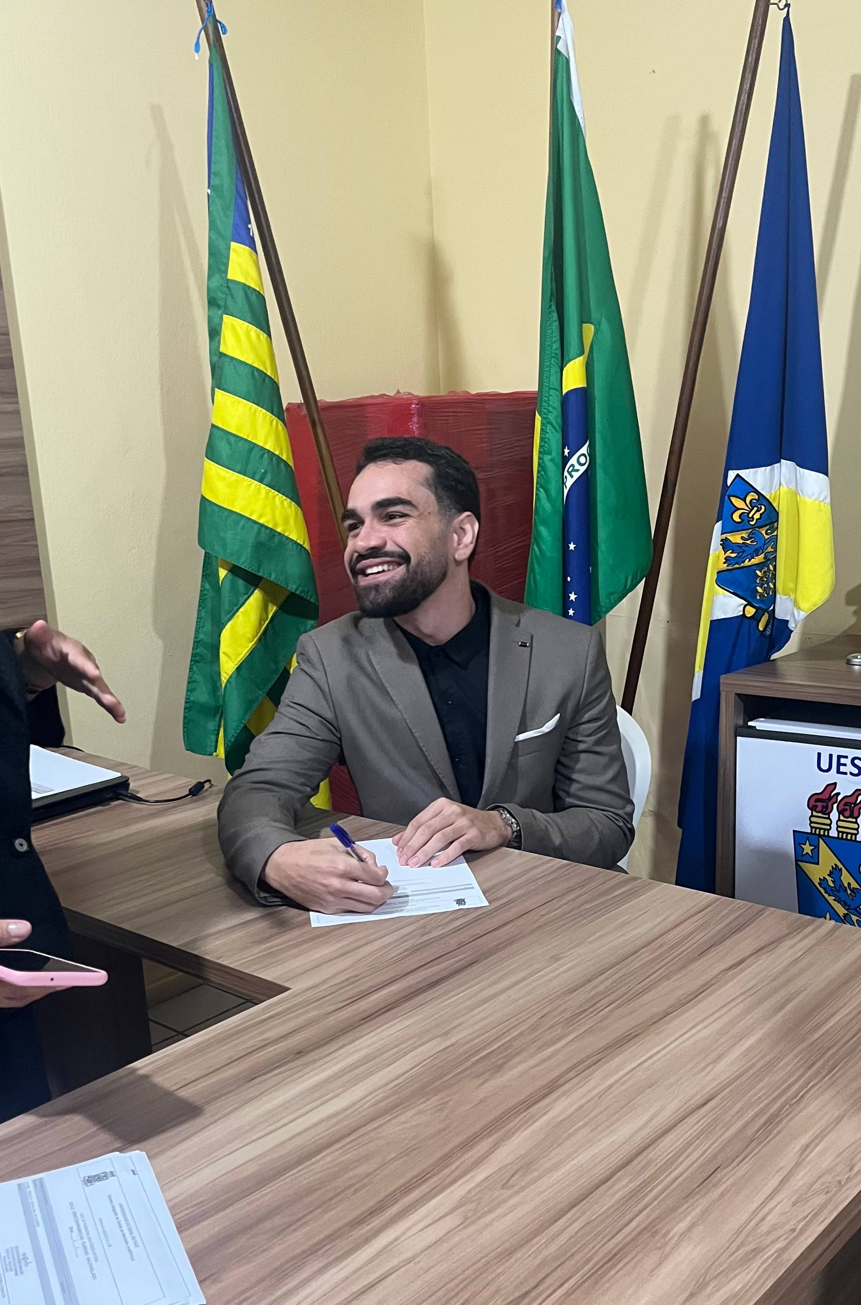 Rickelme Guabiraba sentado à mesa, assinando um documento, com as bandeiras do Piauí, do Brasil e do município ao fundo
