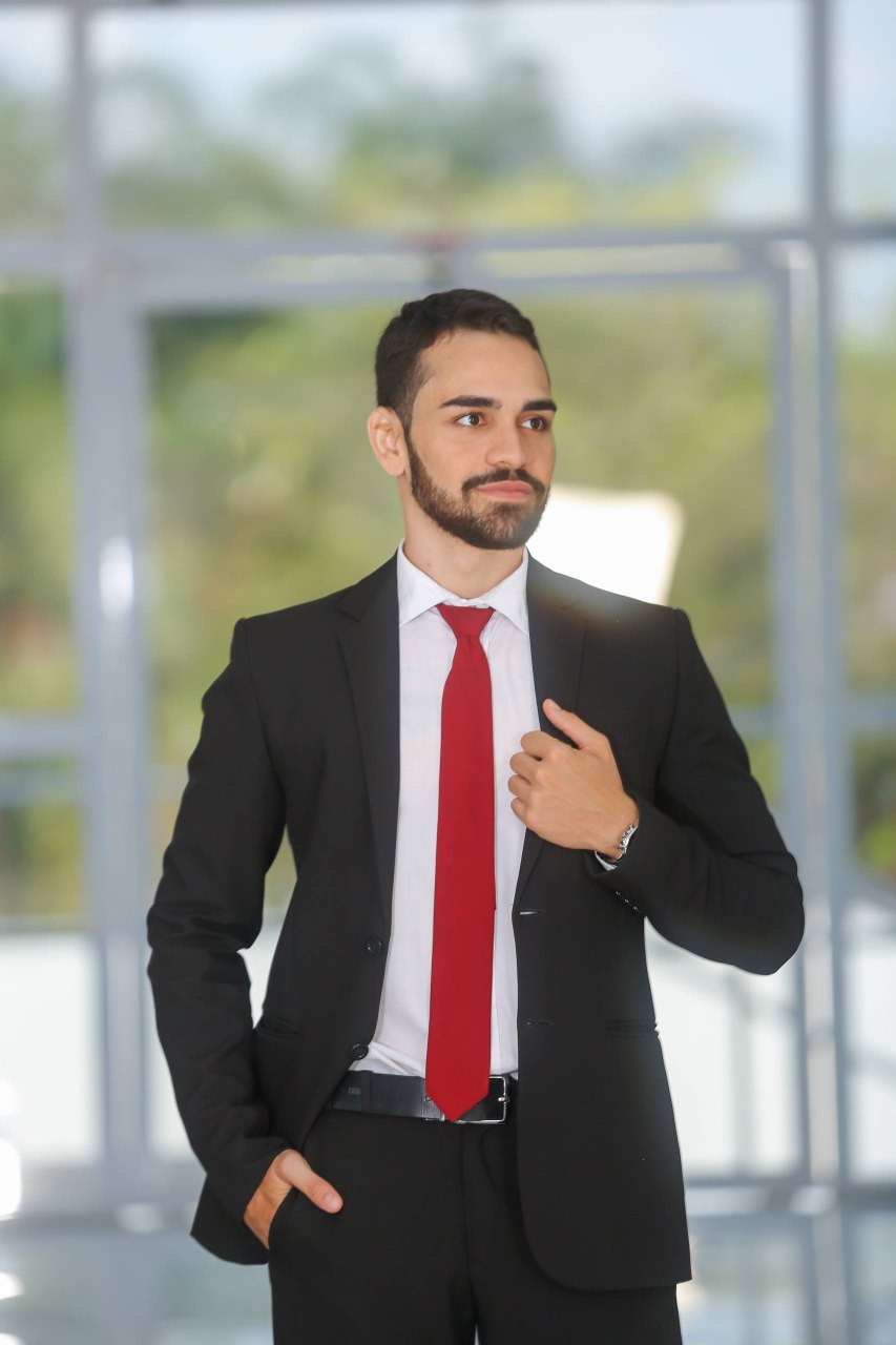 Retrato de corpo inteiro de Rickelme Guabiraba, de terno e gravata, diante de uma fachada de vidro